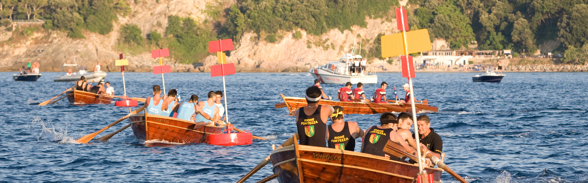 L’immagine mostra una gara di canottaggio a sedile fisso tra quattro squadre, che si svolge in mare aperto in una giornata soleggiata davanti a Porto Santo Stefano In primo piano ci sono quattro barche di legno, ciascuna con un equipaggio di quattro vogatori e una persona al timone o in posizione di comando. Le squadre indossano divise colorate, tra cui si notano maglie nere con scritte gialle ("Rione Fortezza") e maglie celesti ("Rione Valle") Ogni barca ha un paletto verticale con una tavoletta colorata (rossa o gialla), usata per identificare da lontano la boa su cui girare. Sullo sfondo si vedono scogliere e vegetazione, oltre ad alcune barche spettatrici e una motoscafo della sicurezza. L’acqua è calma, e l’atmosfera è quella di una festa e competizione locale, con molta partecipazione. Questa scena richiama una tradizionale regata marina o una gara remiera storica, di Porto Santo Stefano, il Palio Marinaro dell'Argentario.