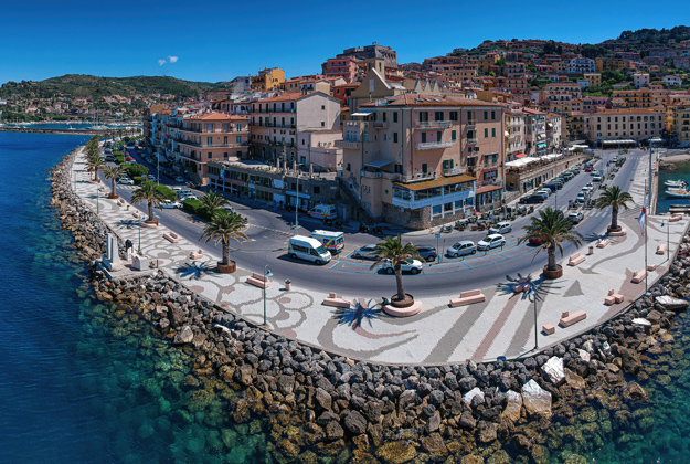 La foto rappresenta il paese di Porto Santo Stefano, vista frontale, scattata da sopra il mare. In primo piano il lungomare con acqua cristallina e sullo sfondo il quartiere storico con la Fortezza Spagnola che sovrasta.