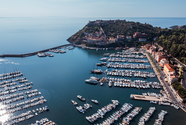La foto rappresenta il porto di Porto Ercole con tutte le sue piccole imbarcazioni, sullo sfondo la parte storica del paese sovrastata dalla Rocca Spagnola