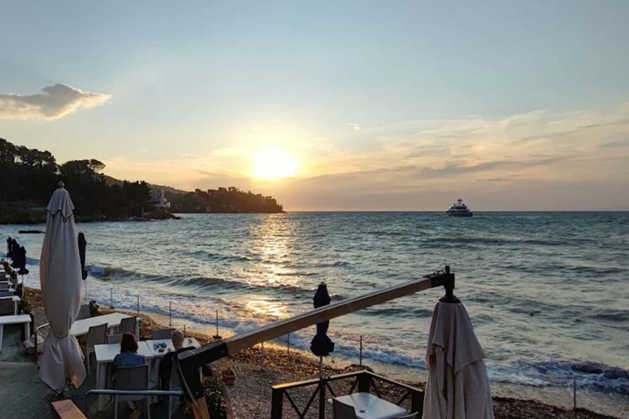 L’immagine ritrae un suggestivo tramonto sul mare, visto da una terrazza panoramica del Ristorante "I due Pini" a la Soda, Argentario. In primo piano, a sinistra, ci sono diversi tavolini apparecchiati con sedie bianche, disposti ordinatamente lungo una passerella che costeggia la riva. Alcuni tavoli sono occupati da persone sedute che godono della vista. Sulla destra, in acqua, si vede un grande yacht bianco ancorato al largo. Il sole è vicino all’orizzonte, in fase di tramonto, e crea un riflesso dorato sulle onde del mare leggermente mosso. A sinistra si staglia la costa collinare, ricoperta da alberi e qualche edificio immerso nel verde. Il cielo è sereno, con qualche nuvola leggera e i colori caldi del tramonto che sfumano dal giallo all’azzurro, rendendo l’atmosfera rilassata e romantica. L’immagine trasmette una sensazione di pace, bellezza naturale e piacevole convivialità, tipica di una serata estiva sul mare.